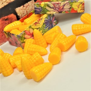 May include: Bright yellow plastic corn kernels scattered on a white surface, with some inside a floral-patterned box. The box has a colorful floral design. The kernels are a vibrant, solid yellow color.