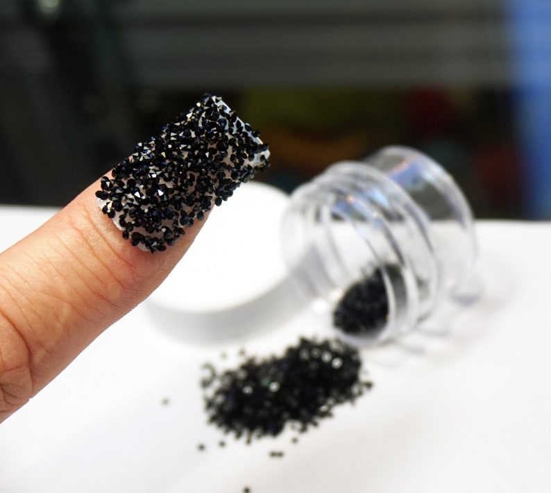 Black nail rhinestones on a finger, with a clear plastic container of black nail rhinestones spilled on a white surface.