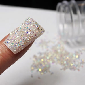 May include: A fingernail decorated with iridescent white micro-beads, with loose beads scattered on a white surface.