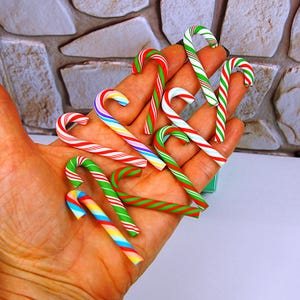 May include: A collection of small, miniature candy cane ornaments featuring various striped patterns in red, green, white, blue, and yellow. These decorative items are shaped like traditional hooked canes and display vibrant, multi-colored diagonal stripes across their smooth surfaces.