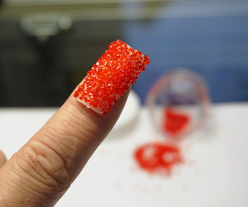 A finger with a red nail art design. The design is made of small, red beads that are clustered together in a rectangular shape. The beads are applied to a clear base coat. The finger is held up against a white background.