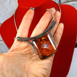 May include: A necklace featuring a large, square, polished amber pendant set in a dark, oxidized silver frame. The pendant is suspended from a thin, flexible silver-toned snake chain that curves around the neck area.