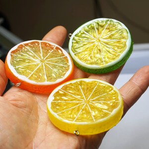 May include: Three colorful, realistic-looking fruit slices made of polymer clay. The slices are a yellow lemon, a green lime, and an orange. Each slice has a white center with a detailed design of the fruit's segments.