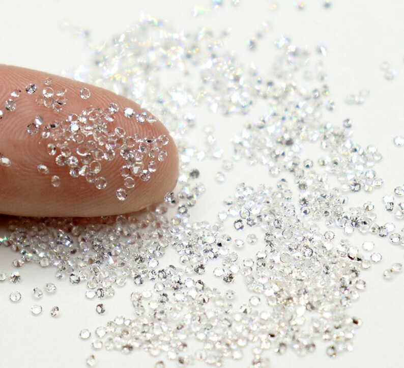 May include: A close-up of a pile of small, clear, round rhinestones. The rhinestones are scattered on a white surface.