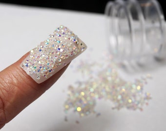 May include: A fingernail decorated with iridescent white micro-beads. The beads are also scattered on a white surface.