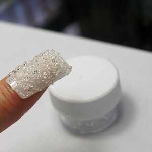 May include: A finger with a white fake nail covered in small, clear rhinestones. A small, clear jar with a white lid is in the background.