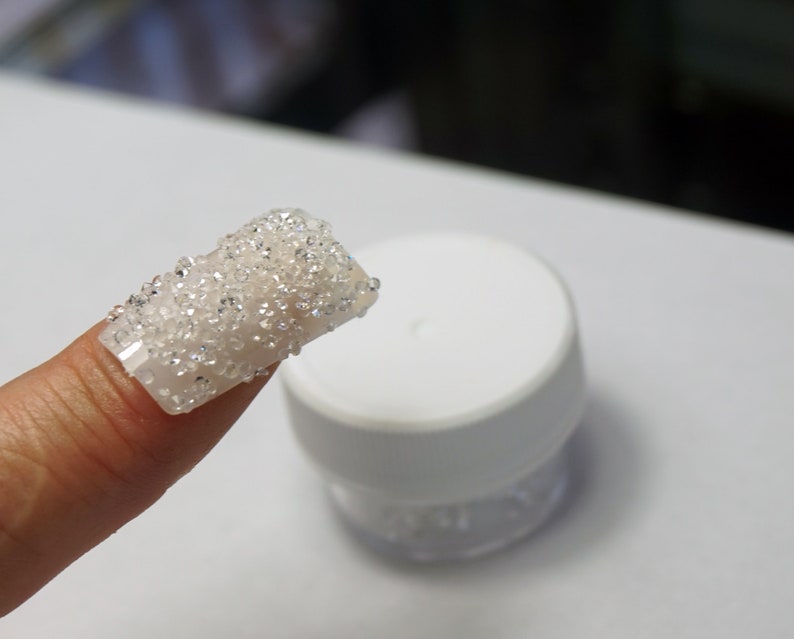 May include: A finger with a white fake nail covered in small, clear rhinestones. A small, clear jar with a white lid is in the background.