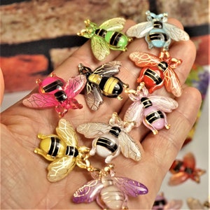 May include: Assortment of colourful bee charms with gold accents. The bees are made of glass and have a variety of colours, including pink, red, green, blue, and purple.