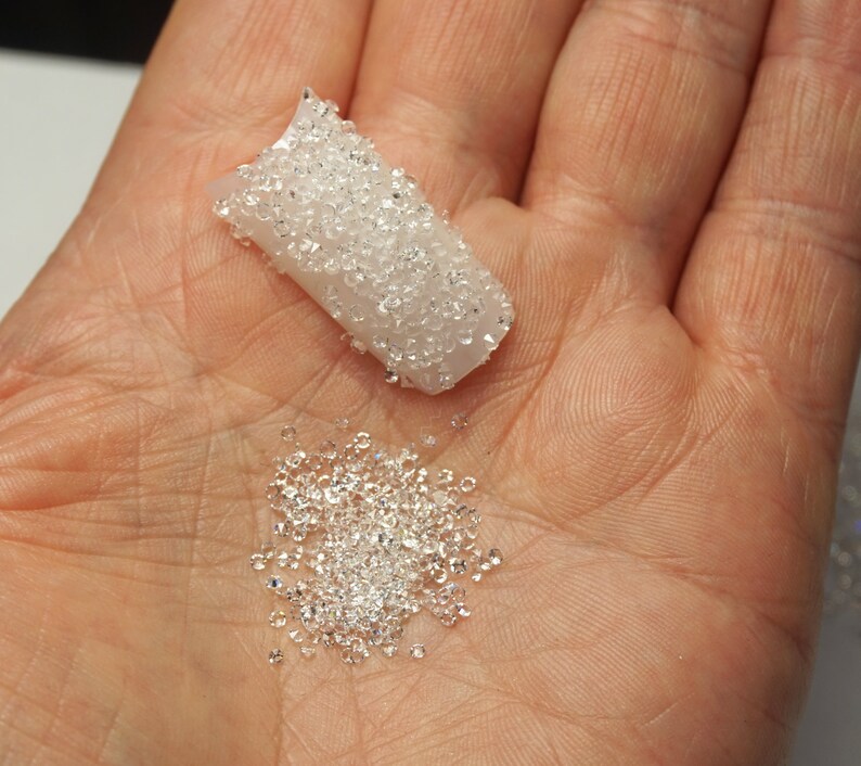 May include: A close-up of a hand holding a small container of clear rhinestones. The container is open and the rhinestones are spilling out onto the hand.