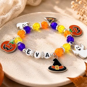 May include: A beaded bracelet featuring alternating yellow, orange, and purple faceted beads, silver-toned spacers, and white letter beads spelling EVA. The bracelet includes metal charms shaped like pumpkins, ghosts, and a black witch hat.