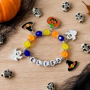 May include: A beaded bracelet featuring the name ELISA in white letter beads, interspersed with yellow, orange, and blue translucent beads. The bracelet includes metal charms shaped like a pumpkin, two ghosts, and two witch hats.