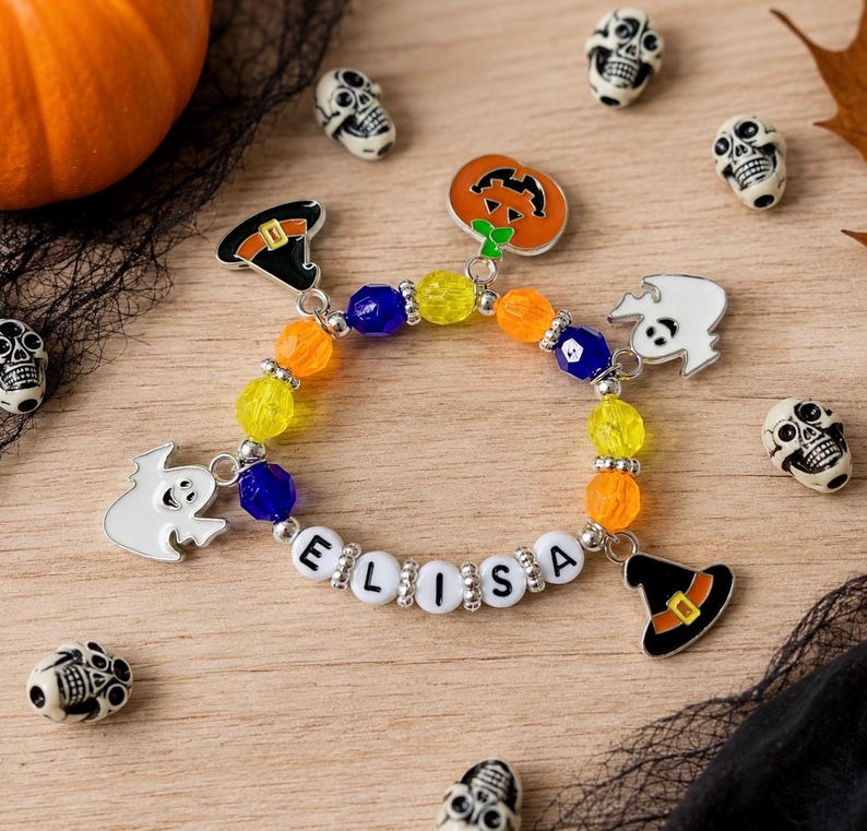 May include: A beaded bracelet featuring the name ELISA in white letter beads, interspersed with yellow, orange, and blue translucent beads. The bracelet includes metal charms shaped like a pumpkin, two ghosts, and two witch hats.