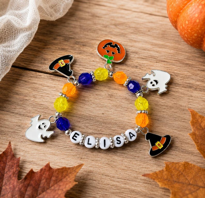 May include: A beaded bracelet featuring colorful translucent yellow, orange, and blue beads, silver spacers, and white letter beads spelling ELISA. The bracelet includes metal charms of a pumpkin, two ghosts, and two witch hats.