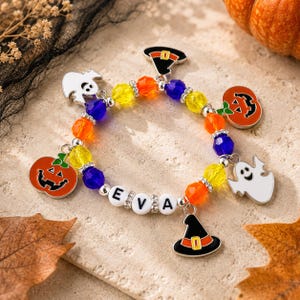May include: A beaded bracelet featuring colorful yellow, orange, and blue faceted beads, silver-toned spacers, and white letter beads spelling EVA. The bracelet includes dangling charms shaped like white ghosts, orange pumpkins, and black witch hats.