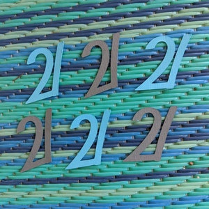 May include: Six paper cutouts of the number 21, three in blue and three in gray, arranged in a grid pattern on a blue and green striped background.