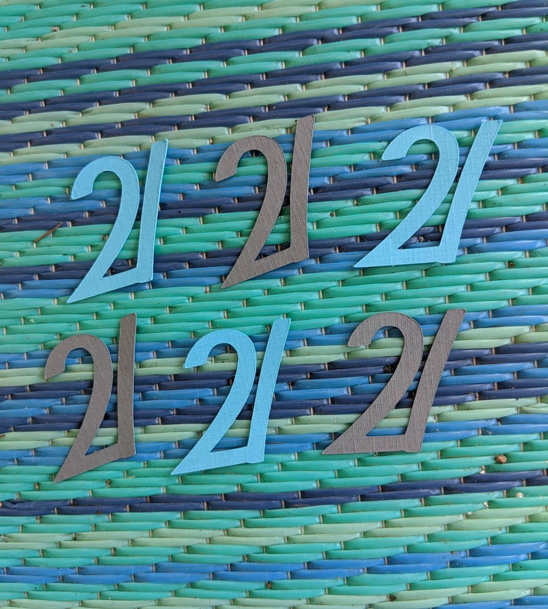 May include: Six paper cutouts of the number 21, three in blue and three in gray, arranged in a grid pattern on a blue and green striped background.