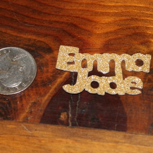 May include: Gold glitter letters spelling out the name "Emma Jade" on a wooden surface.