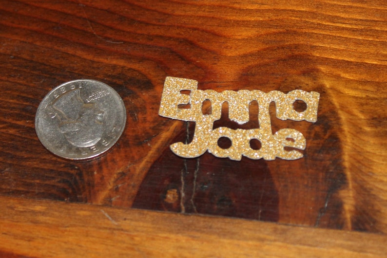 May include: Gold glitter letters spelling out the name "Emma Jade" on a wooden surface.