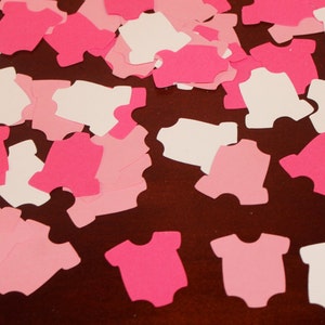 May include: Pink, white and light pink paper cut-outs of baby onesies. The onesies are scattered on a brown surface.