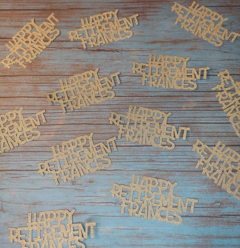 K&ouml;nnte beinhalten: Goldene Konfetti mit Glitzer und dem Text "Happy Retirement Frances" auf einer blauen Holzoberfl&auml;che verstreut.
