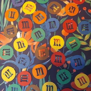 May include: A collection of colorful paper circles with the letter "M" cut out of the center. The circles are arranged in a random pattern on a blue background with floral designs.