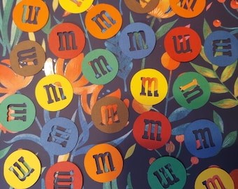 May include: A collection of colorful paper circles with the letter "M" cut out of the center. The circles are arranged in a random pattern on a blue background with floral designs.