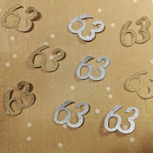May include: A collection of gold and silver glitter confetti shaped like the number 63. The confetti is scattered on a brown background with white polka dots.