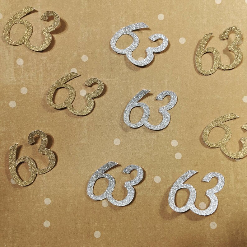 May include: A collection of gold and silver glitter confetti shaped like the number 63. The confetti is scattered on a brown background with white polka dots.