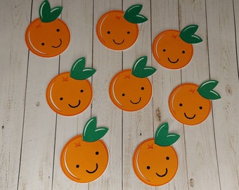 May include: Eight orange felt cutouts of oranges with green leaves and smiling faces. The oranges are arranged in a circle on a light wood background.