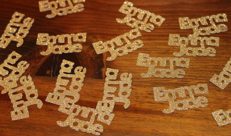 May include: A collection of gold glitter paper cutouts with the name "Emma Jade" written on each one.