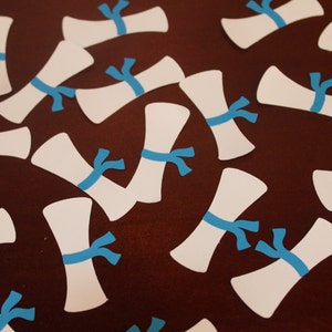May include: A collection of white paper cutouts shaped like diplomas with blue ribbon accents. The cutouts are scattered on a brown surface.