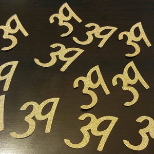 May include: A collection of gold glitter-covered paper cutouts of the number 39. The cutouts are scattered on a dark brown surface.