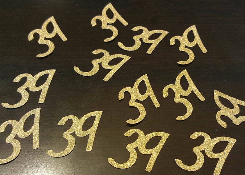 May include: A collection of gold glitter-covered paper cutouts of the number 39. The cutouts are scattered on a dark brown surface.