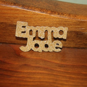May include: Gold glitter letters spelling out the name "Emma Jade" on a brown wooden surface.
