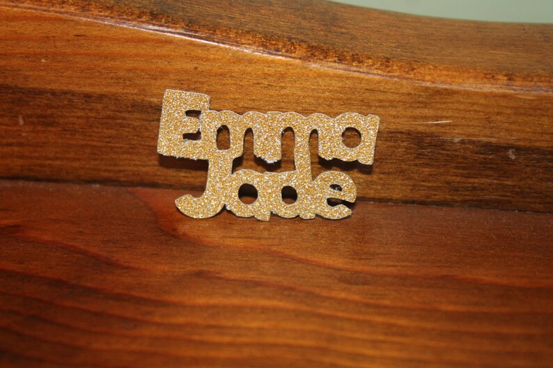 May include: Gold glitter letters spelling out the name "Emma Jade" on a brown wooden surface.