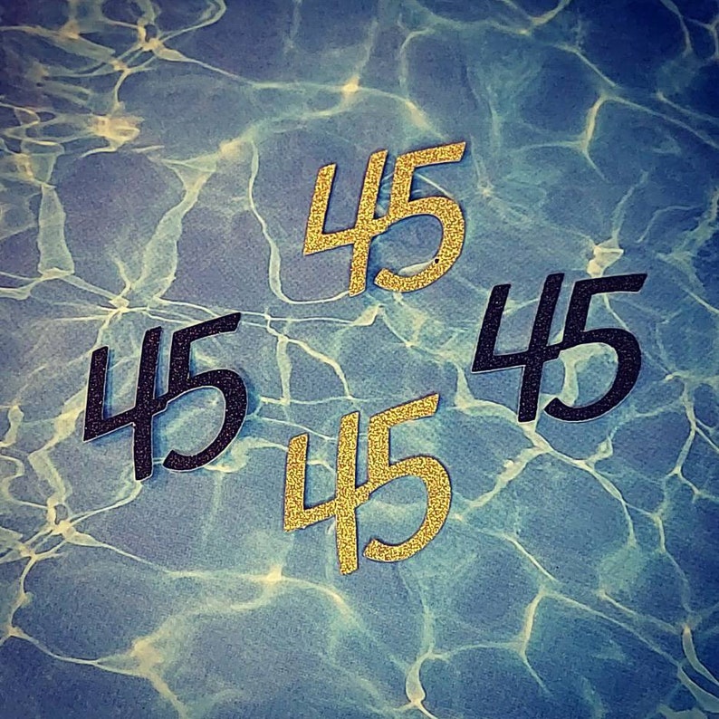 May include: Four black and gold paper cutouts of the number 45. The cutouts are arranged in a diamond shape on a blue and white background.