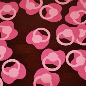 May include: Pink paper cutouts of baby pacifiers scattered on a dark brown surface.