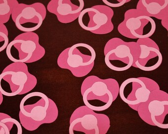 May include: Pink paper cutouts of baby pacifiers scattered on a dark brown surface.