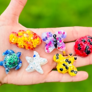 May include: A collection of colorful, handcrafted resin charms in various shapes, including a fish, butterfly, ladybug, turtle, starfish, and another butterfly. The charms are filled with small, colorful beads and set against a blurred green background.