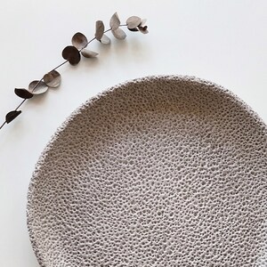 May include: A large, round, grey ceramic plate with a textured surface. The plate is on a white background.