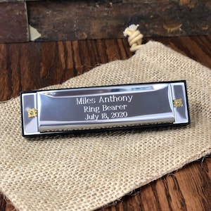 May include: A rectangular silver-toned harmonica with black trim and gold-colored side screws. The metal surface is engraved with the text: Miles Anthony, Ring Bearer, July 18, 2020. The instrument rests on a tan, textured burlap drawstring pouch.