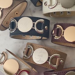 May include: A collection of textured leather bracelets in various colors including black, white, brown, and burgundy. Each bracelet features a central circular metal disc, some engraved with letters or the text Serve with Love, and metallic hardware.