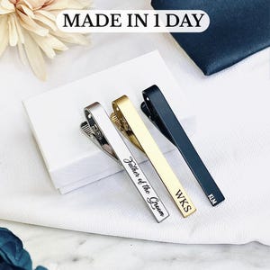 May include: Three personalized tie clips in silver, gold, and black, each engraved with initials or text. The gold clip reads "Father of the Groom." The image includes the text "MADE IN 1 DAY."