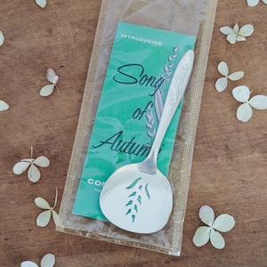 May include: A silver serving spoon with a wheat stalk design, packaged in a clear plastic sleeve with a green card that reads "Song of Autumn". The spoon has a slotted design and is surrounded by white flower petals.