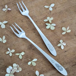 May include: Two silver forks with three tines each, on a wooden surface. The handles have an engraved design. Small, dried, light green and white flowers are scattered around the forks, creating a rustic aesthetic.