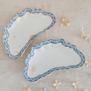 May include: Two white, scalloped-edge serving dishes with a blue and gold decorative border. The dishes are placed on a neutral-colored linen surface, with dried flowers scattered around them. The dishes have a unique, elegant design.