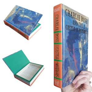 May include: A hardcover book titled "Charlie Bone and the Castle of Mirrors" by Jenny Nimmo. The cover features a blue background with a boy's portrait. The book has an orange spine with the title and author's name. The book is open, revealing a hollowed-out interior.