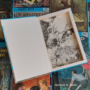 May include: A collection of vintage Hardy Boys mystery books with blue covers. The books are arranged in a stack with the titles visible. The top book is open to reveal a black and white illustration of a shadowy figure.