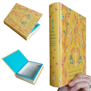May include: A hardcover book titled The Secret Garden by Frances Hodgson Burnett with a yellow cover featuring pink floral patterns, blue birds, and a white key. The book is hollowed out to create a hidden compartment with a bright blue interior lining.