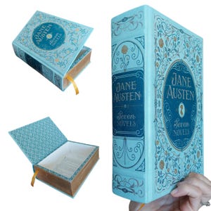May include: A light blue book-shaped storage box titled Jane Austen Seven Novels. The exterior features intricate gold and dark blue floral filigree patterns. The box opens to reveal a hollow interior with a patterned lining and a yellow ribbon bookmark.